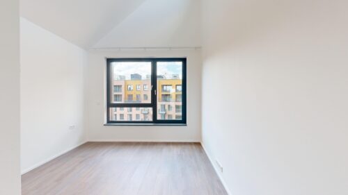 Empty white-walled room with light wood flooring and a large black-framed window overlooking a residential building exterior.