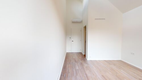 Bright empty hallway/room with white walls, light wood floor, wall-mounted air conditioner, and a door at the far end.