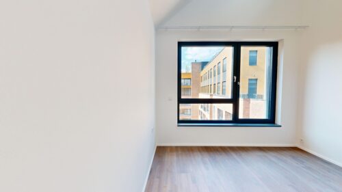 Empty white-walled room with light wooden floor and a large black-framed window showing a brick apartment building outside.