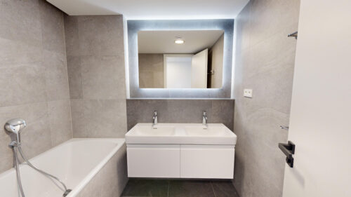Modern bathroom with grey tiled walls, white double sink vanity, and backlit rectangular mirror above it, door open in background