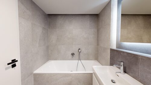 Modern gray-tiled bathroom with a built-in white tub, wall-mounted faucet, and handheld shower head, in a minimalist design.