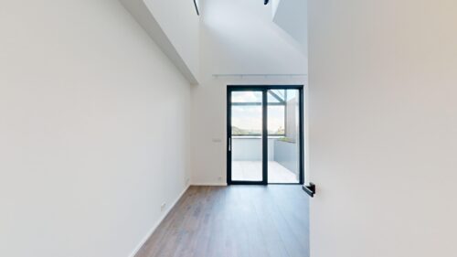 Bright minimalist hallway with white walls and light wood flooring, leading to a glass-framed door opening to a balcony with a blue sky.