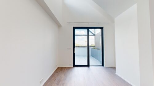 Bright empty room with white walls, light wood floor, and a glass door opening to a balcony with blue sky outside.