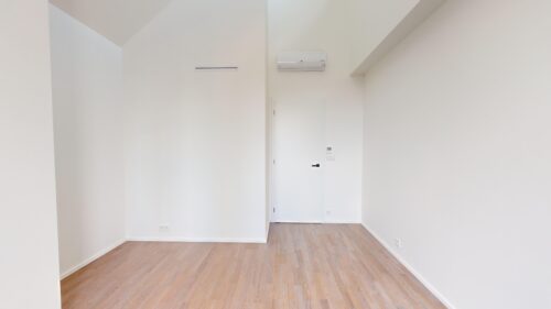 Bright empty room with white walls, light wood floor, a wall-mounted air conditioner, and a closed door nearby.
