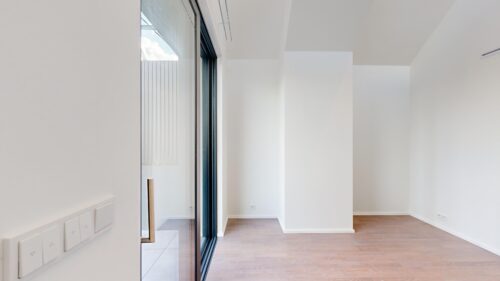Bright minimalist interior with white walls, tall ceiling, and a glass sliding door revealing an outdoor area with blue sky and metal fence.