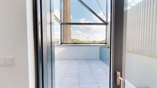 Open glass door to a tiled balcony with a metal railing; distant chimney and cityscape under blue sky.