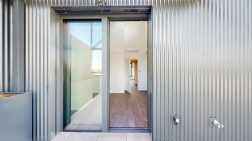 Metal-clad exterior with a glass door opening into a white interior hallway and hardwood floor seen through the doorway