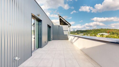 Modern rooftop terrace with corrugated gray metal walls, glass doors, and large tiled floor under a blue sky.