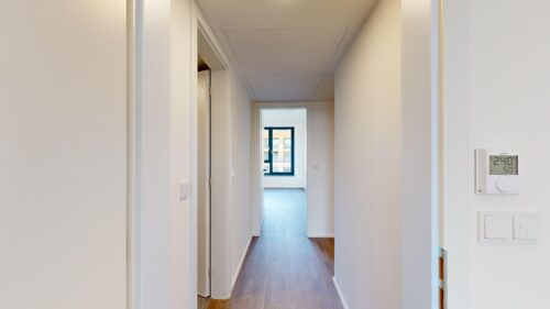 Bright white hallway with a wooden floor, door frames on the left, and a blue-framed window at the far end; a digital thermostat on the right wall shows 24°C.
