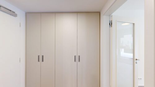 Tall beige built-in wardrobe with four doors and silver handles in a bright hallway beside a white-framed glass door