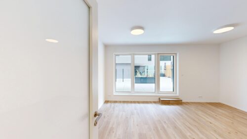 Bright empty room with white walls, light wood floor, a large window showing an outdoor courtyard, and a door opening on the left.