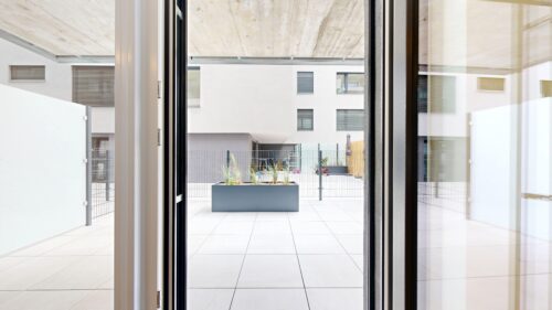 Interior view through glass doors opening to a modern courtyard with a rectangular planter and white building exterior in the background, including windows and a fence.