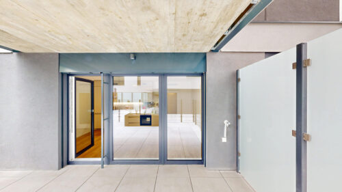 Modern building entrance with open glass doors, gray framing, and a bright interior visible beyond.