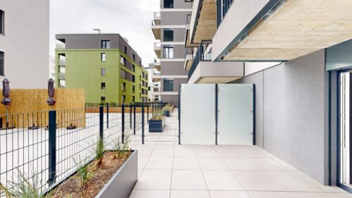 Wide view of a modern residential courtyard with tiled terrace, black metal fence, and frosted glass panels between buildings.