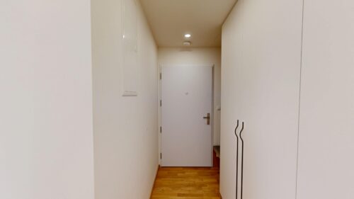 Narrow white hallway with a wooden floor and a white door at the far end, flanked by tall white cabinets on the right.