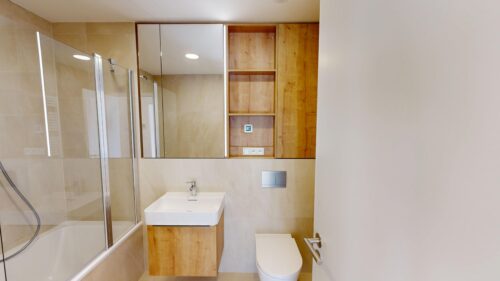 Modern bathroom with a glass-enclosed shower, beige tiles, a white wall-mounted sink, and wooden storage cabinets alongside a wall-hung toilet.
