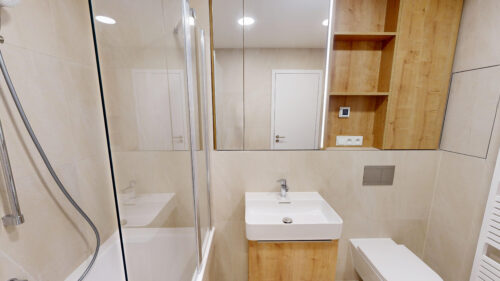 Modern bathroom with glass shower, white rectangular sink, and wooden shelves on beige tiled walls.