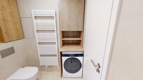 Bathroom with a white towel radiator, wooden storage shelves, and a front-loading washing machine between beige tiled walls and a white door.