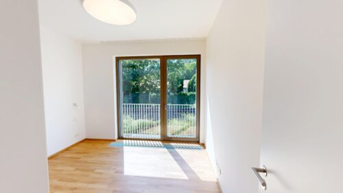 Bright empty room with wooden floor and a large glass door leading to a balcony, greenery outside.