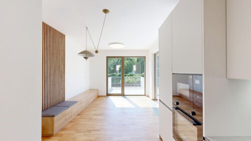 Bright modern kitchen with built-in ovens on the right, wooden bench seating, and glass doors to a balcony overlooking greenery.