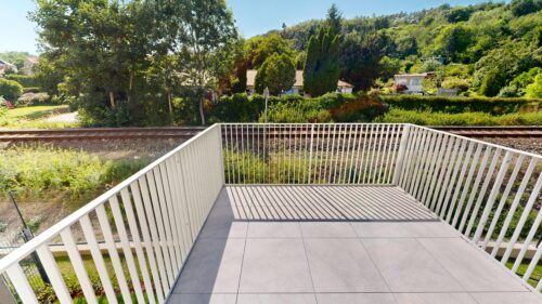 Empty balcony with white railing and large tiled floor, overlooking railway tracks and a green garden with trees and houses in the distance.