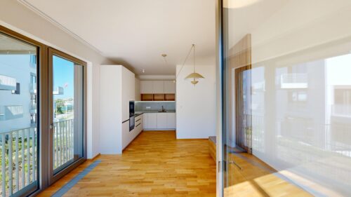 Bright open-concept kitchen with white cabinets, a gray backsplash, and warm wooden floors, seen through large sliding glass doors to a balcony.