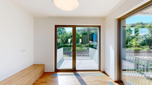 Bright, modern interior with a large glass door opening to a balcony and green outdoor scenery beyond the railing, sunlight flooding the room.