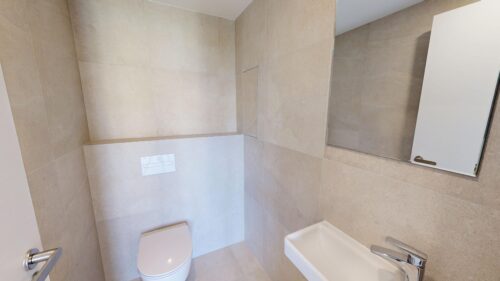 Beige tiled bathroom with wall-mounted toilet and white sink seen from a corner, mirror on the wall, and a door handle visible.