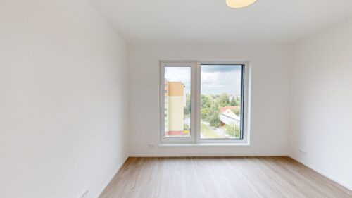Empty white-walled room with light wood flooring and a large window revealing a yellow apartment building and trees outside, bathed in natural light.