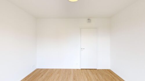Plain white room with light wood floor, a closed white door centered on the back wall, and a small air vent above the door.