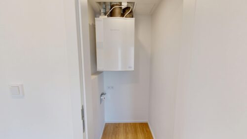 Narrow utility closet with a wall-mounted white boiler and exposed pipes above it on a white wall. A wooden floor is visible below.