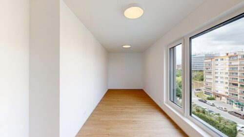 Empty modern room with white walls, light wood floor, and a large corner window overlooking city buildings outside