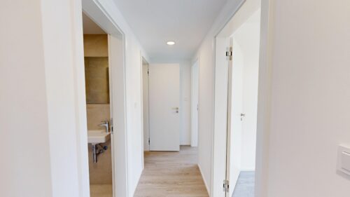 Bright white hallway with multiple open doors along a light wood floor, leading to a door at the end; a bathroom sink is visible through the left doorway.