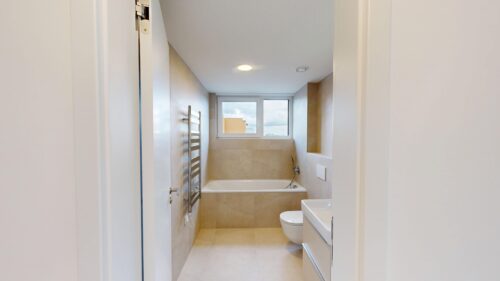 Bright, modern bathroom with beige tiled walls, a built-in tub along the far wall, and a wall-mounted chrome towel warmer under/near the window.