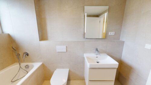 Modern beige-tiled bathroom with a white bathtub, handheld shower, wall-hung toilet, and a white rectangular sink under a mirror.