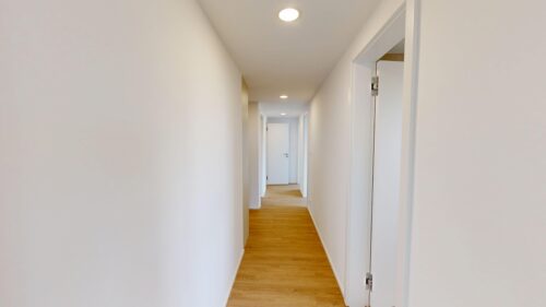 Bright narrow hallway with white walls, light wood floor, and doors along both sides under recessed ceiling lights at the end.
