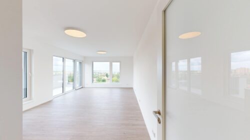 Bright, empty living space with white walls, pale wood flooring, and large windows opening to a balcony with a city view
