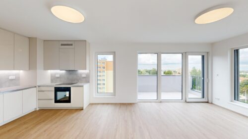 Bright, open-plan kitchen with white cabinetry, built-in oven, and gray backsplash beneath under-cabinet lighting; large windows and balcony doors overlook a cityscape.