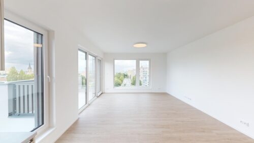 Bright, empty living space with white walls, large windows and a balcony overlooking a cityscape; light wood flooring.