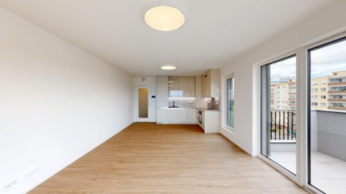 Bright, open-plan living area with white walls, light wood floors, and a modern kitchen along the back wall; large glass doors to a balcony on the right.