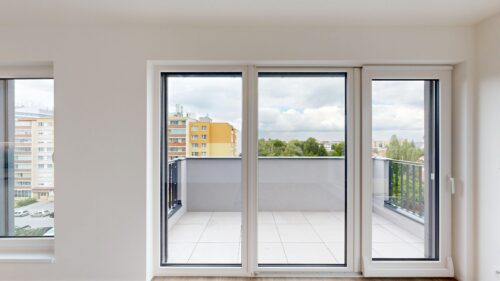 Interior room with large glass sliding doors opening to a balcony, overlooking a cityscape with apartment buildings and trees.